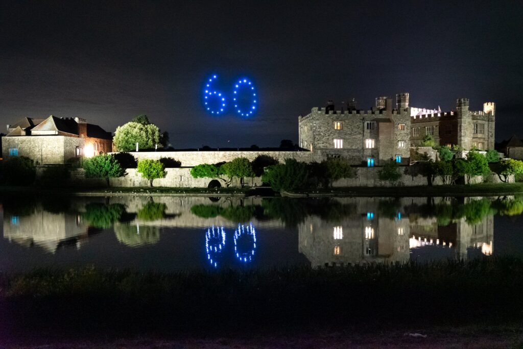 A castle at night with “60” formed by blue lights in the sky and reflected in water below.