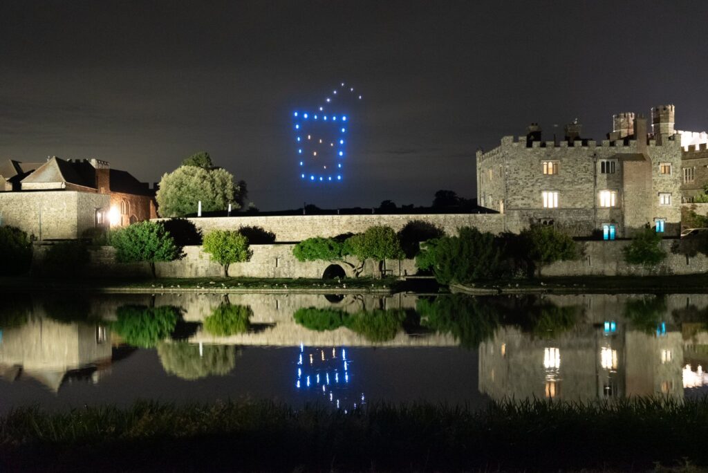 A night scene with a drone light display forming a blue crown over a castle, reflected in a lake.