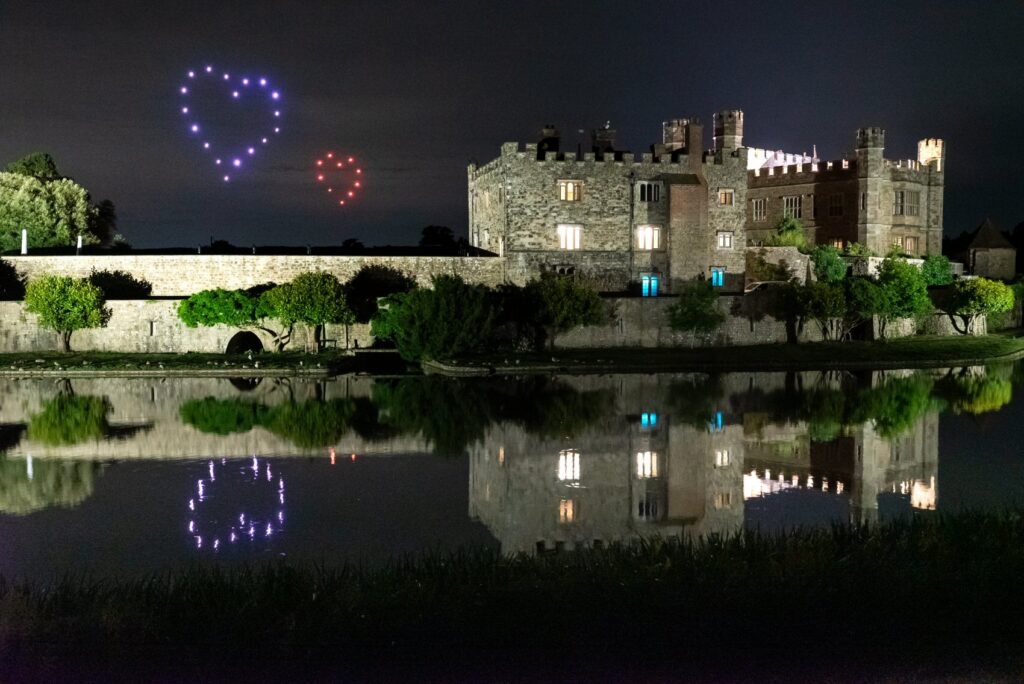 A castle by a lake at night with heart-shaped lights in the sky and their reflections in the water.