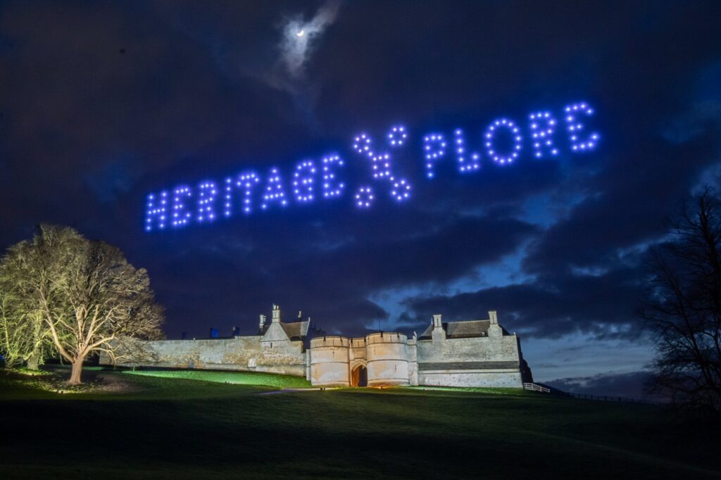 A castle at night with “HERITAGE XPLORE” spelled out in blue lights by drones in the sky above.