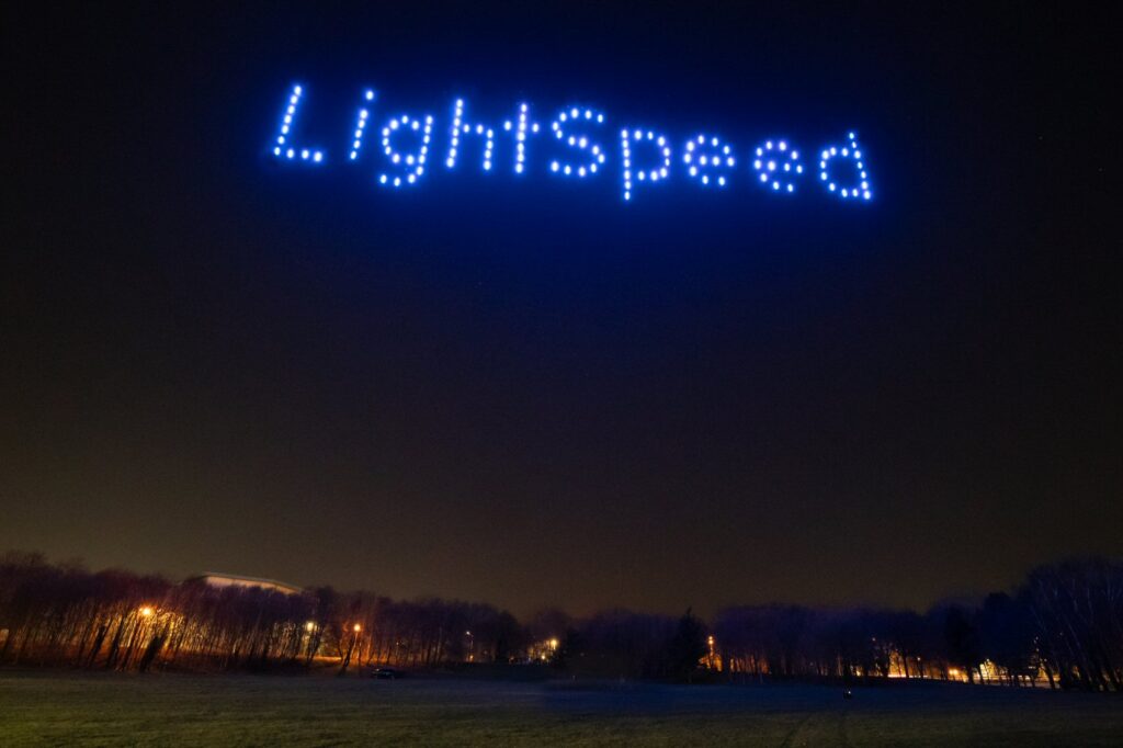 LightSpeed" spelled out in blue lights by drones over a dark, open field at night.