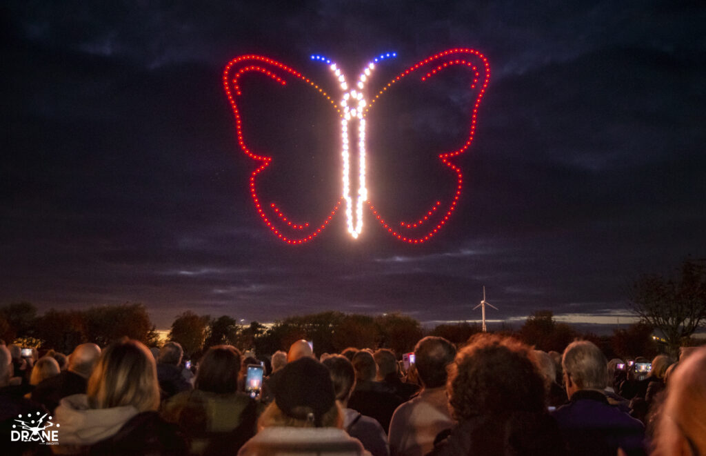 A crowd watches drones form a glowing butterfly shape in the night sky.